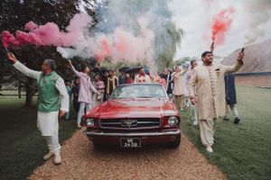 A classic red car surrounded by celebrants with colorful smoke effects at an outdoor event.
