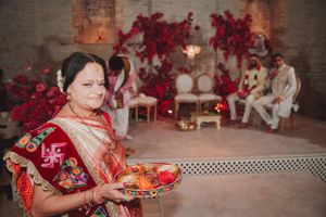 A person in traditional red Hindu attire holds a ceremonial plate surrounded by vibrant red decorations at a cultural event.