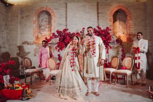 Traditional Indian wedding ceremony with red flower arch and brick wall backdrop.