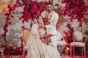 Wedding couple poses under red floral arch during traditional Indian celebration.