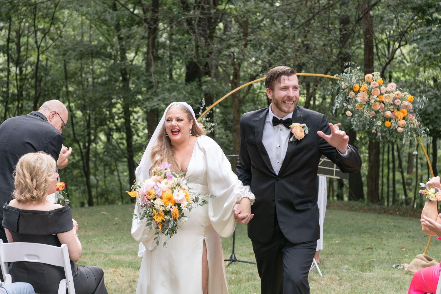 Happy couple exits wedding ceremony under floral arch carrying bright orange and yellow bouquet.