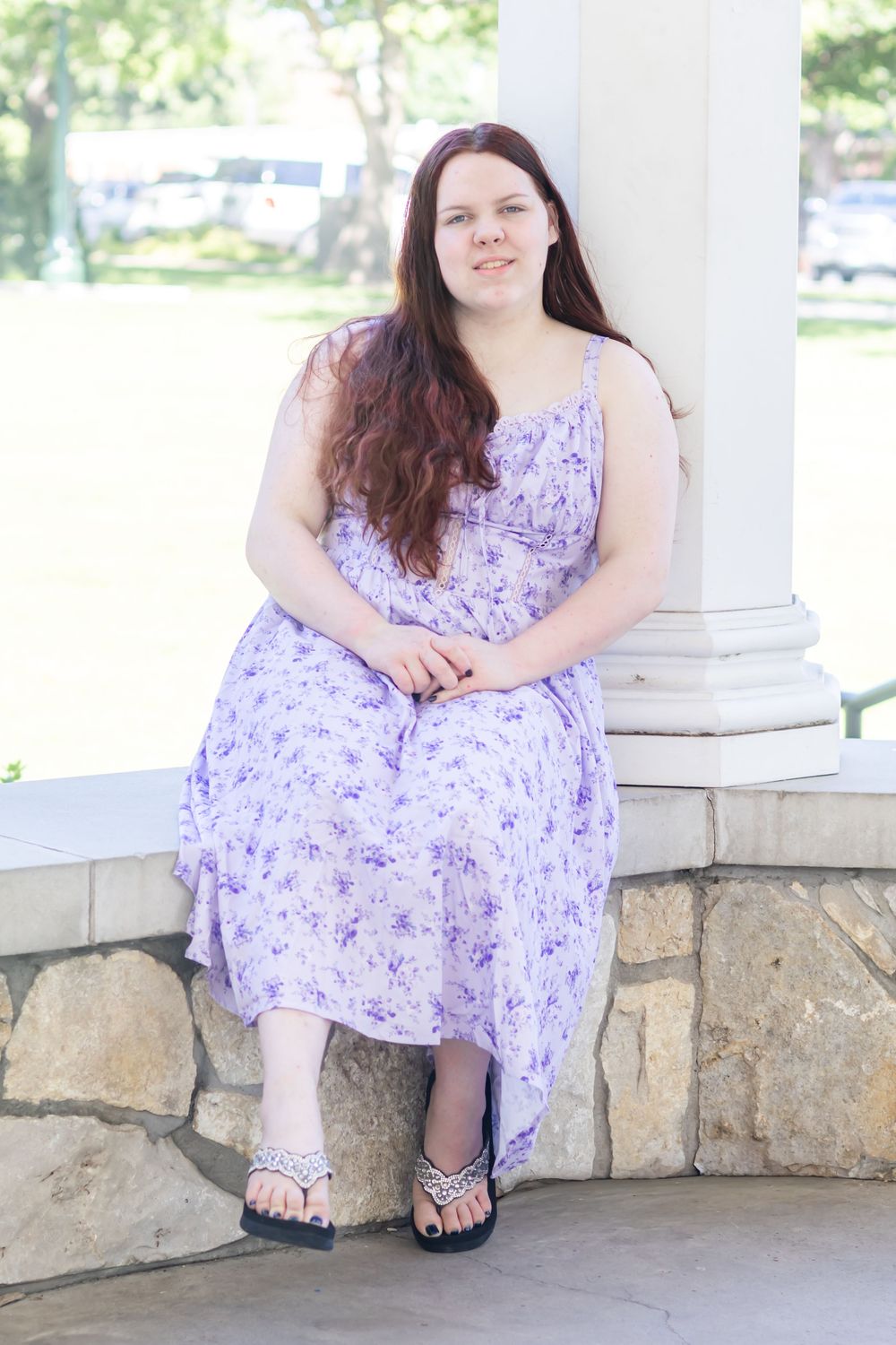 Young woman with long red hair wearing a purple floral sundress and sandals, sitting by a stone pillar outdoors.