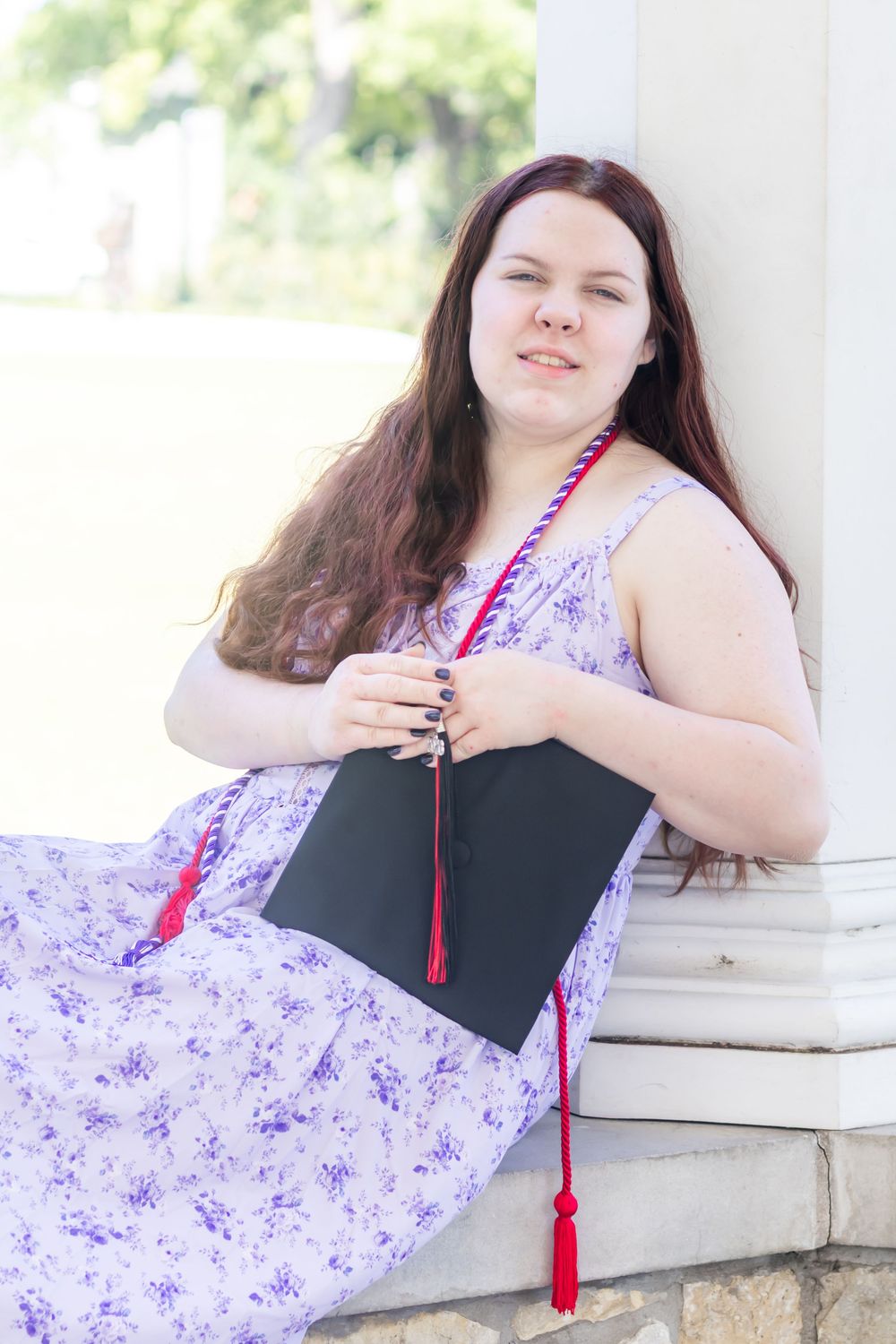Young woman in floral dress holding graduation cap with red tassel, leaning against white column outdoors.