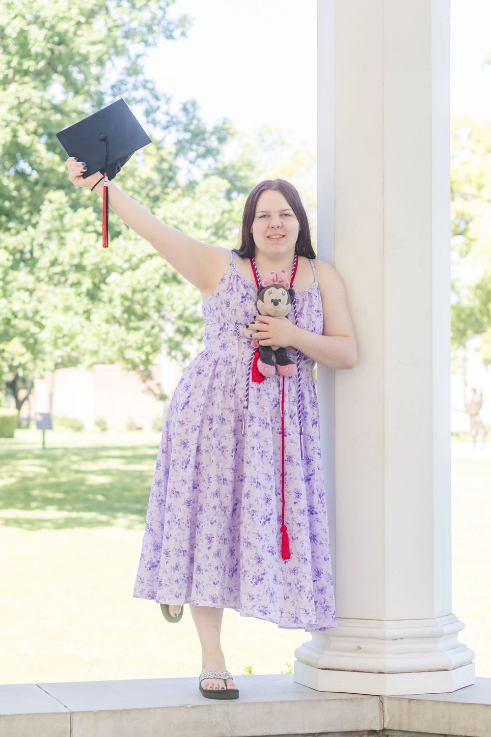 Woman in purple floral dress holding graduation cap and stuffed animal, celebrating outdoors near white column.