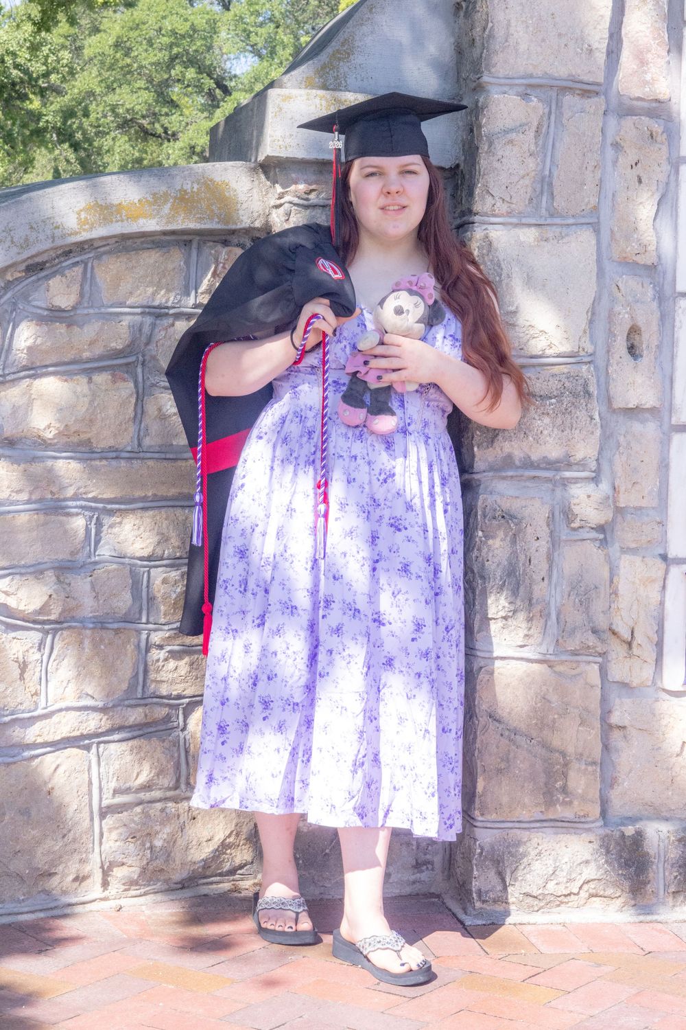 Young woman in graduation cap and gown holding a stuffed animal, posing against a stone wall outdoors.