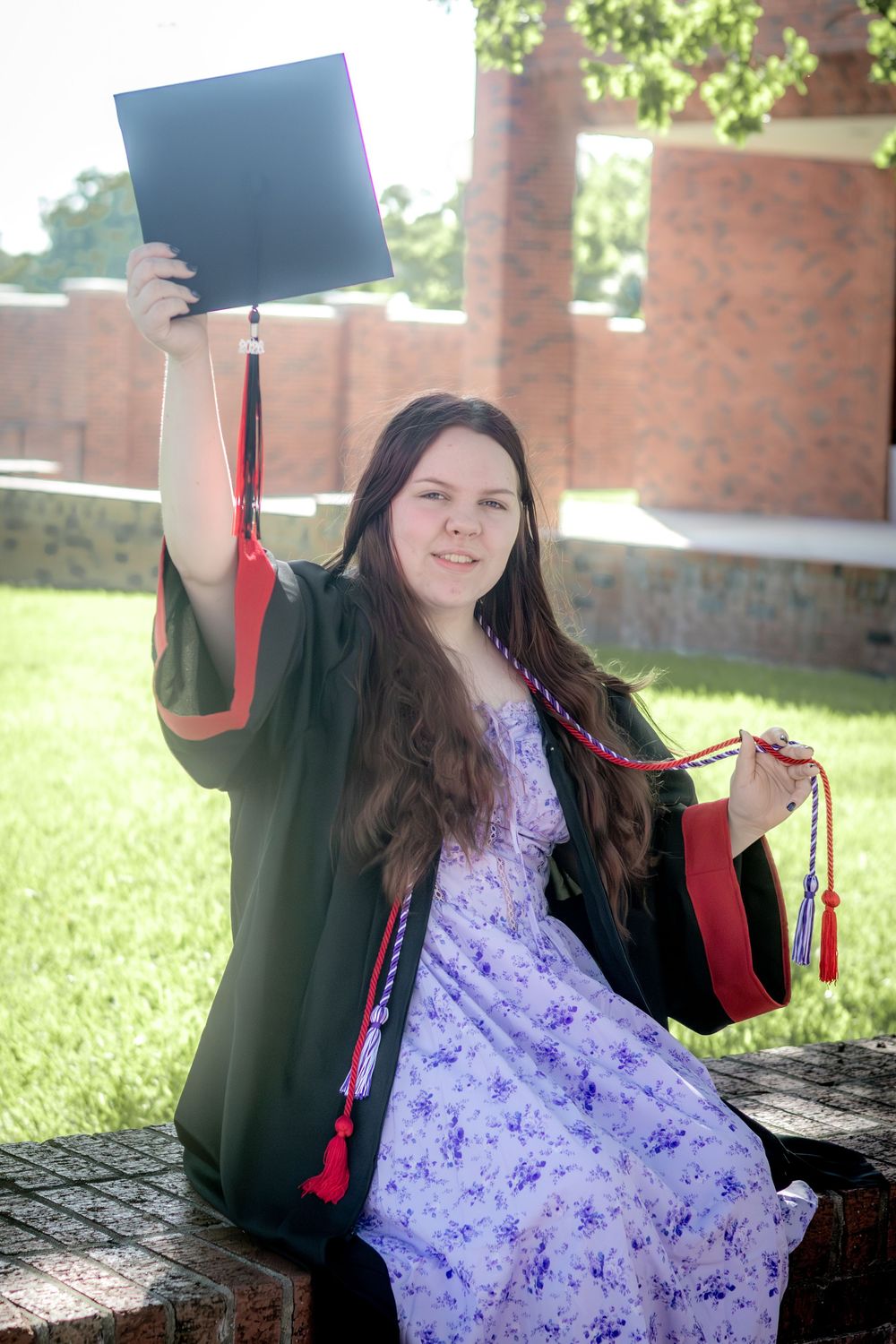 Young woman in graduation gown holding cap and diploma outdoors on sunny campus grounds.