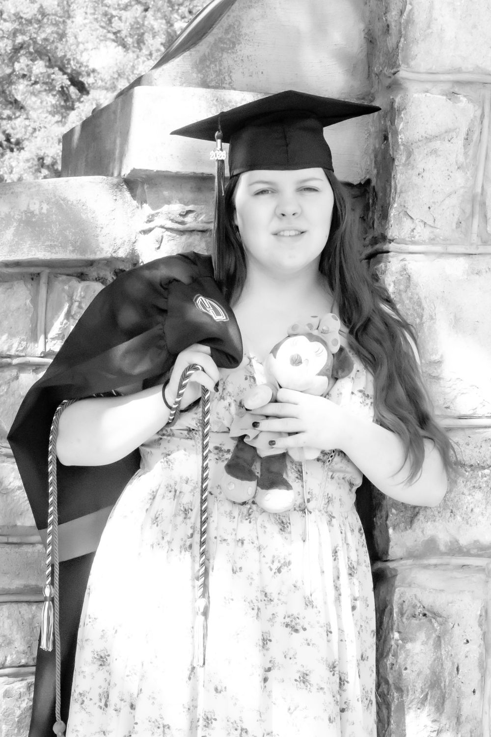 Black and white photo of a young woman in graduation cap holding flowers and gown, posing by stone wall.