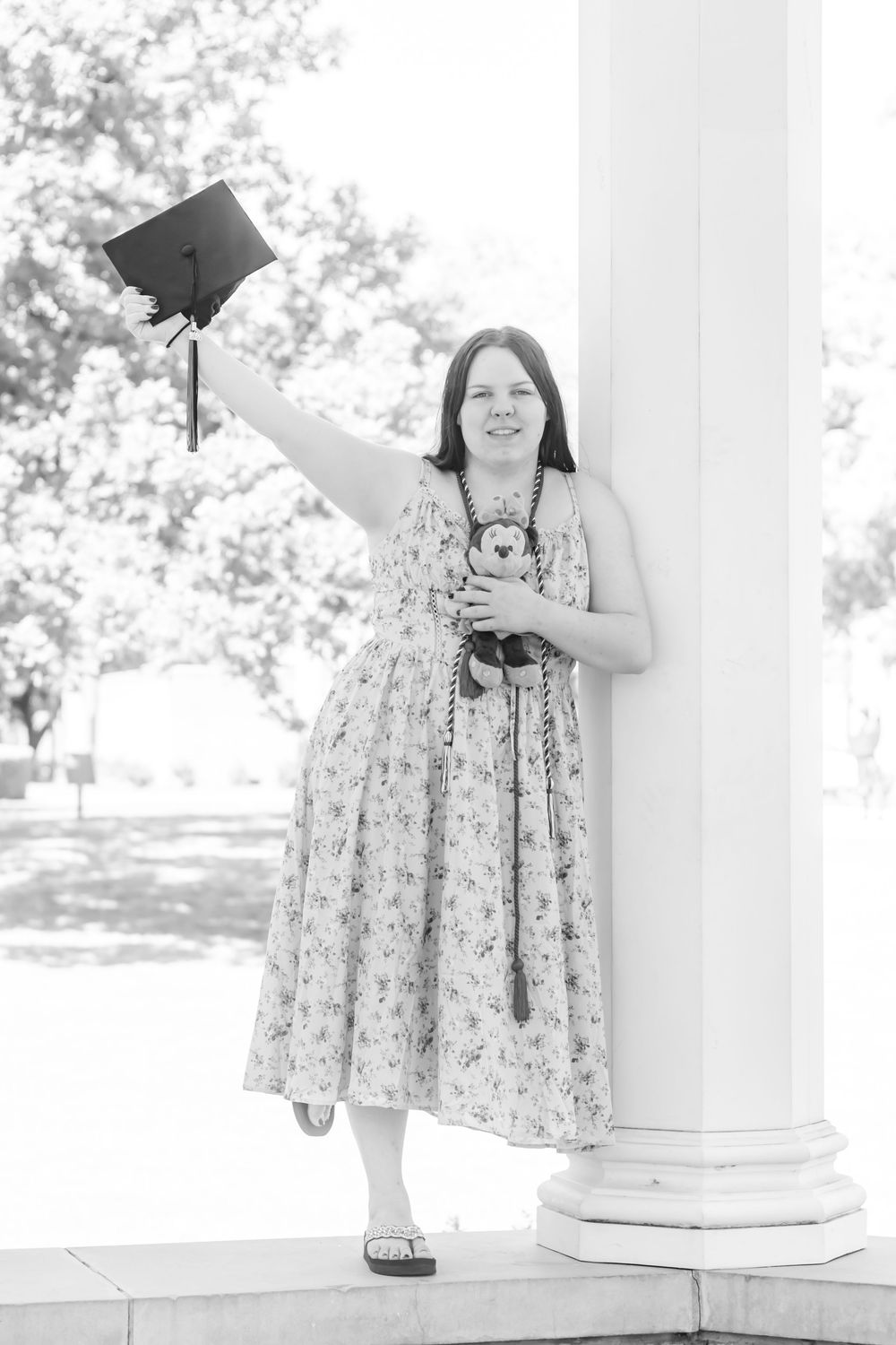 Young woman in floral dress holding graduation cap aloft beside white column, celebrating academic achievement outdoors.