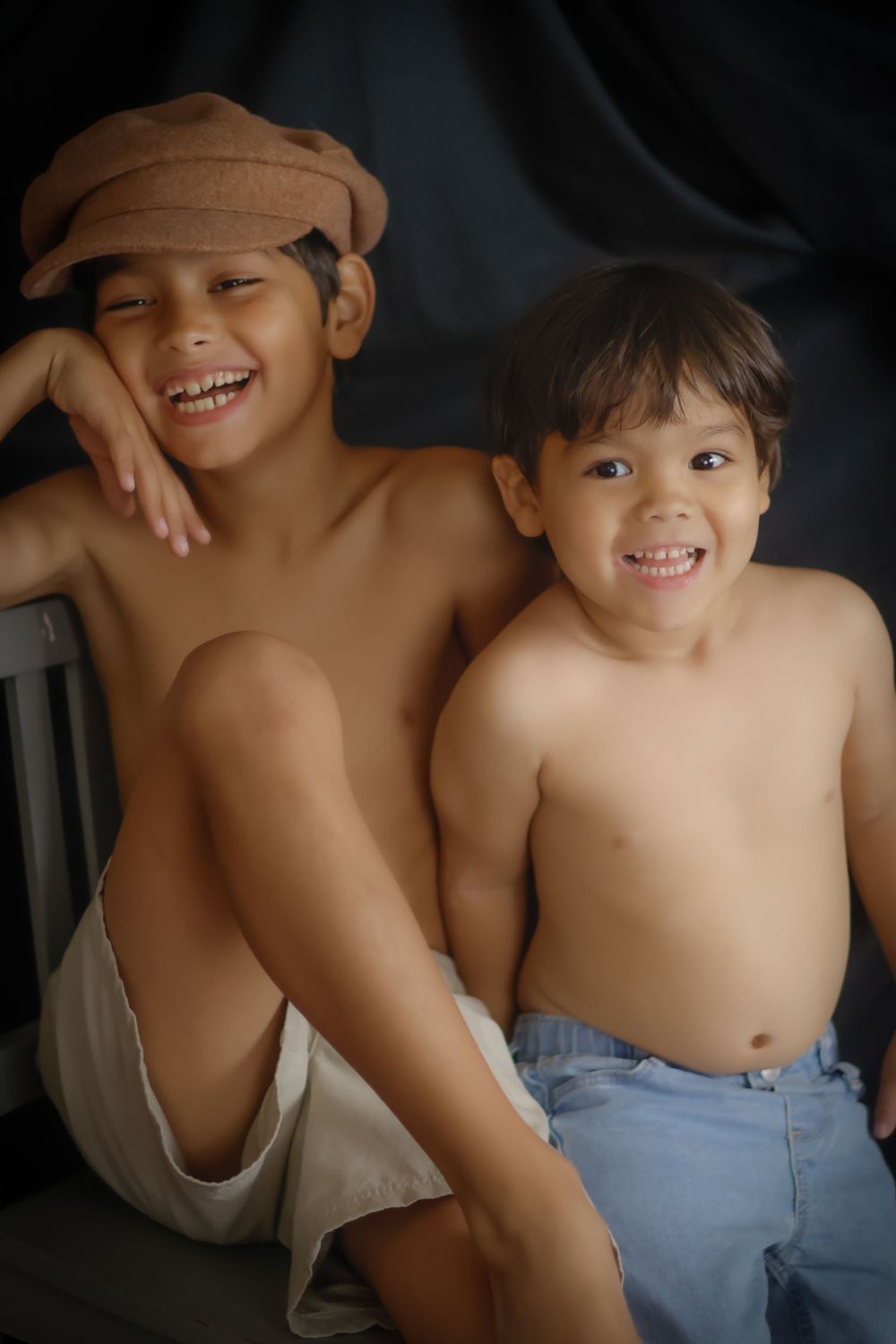 Two smiling shirtless boys, one wearing a brown cap, pose together against a dark background.