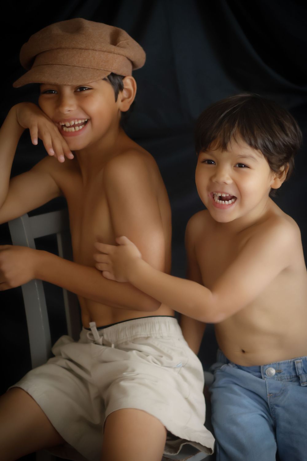 Two smiling shirtless boys posing together, one wearing a brown newsboy cap, against a dark background.