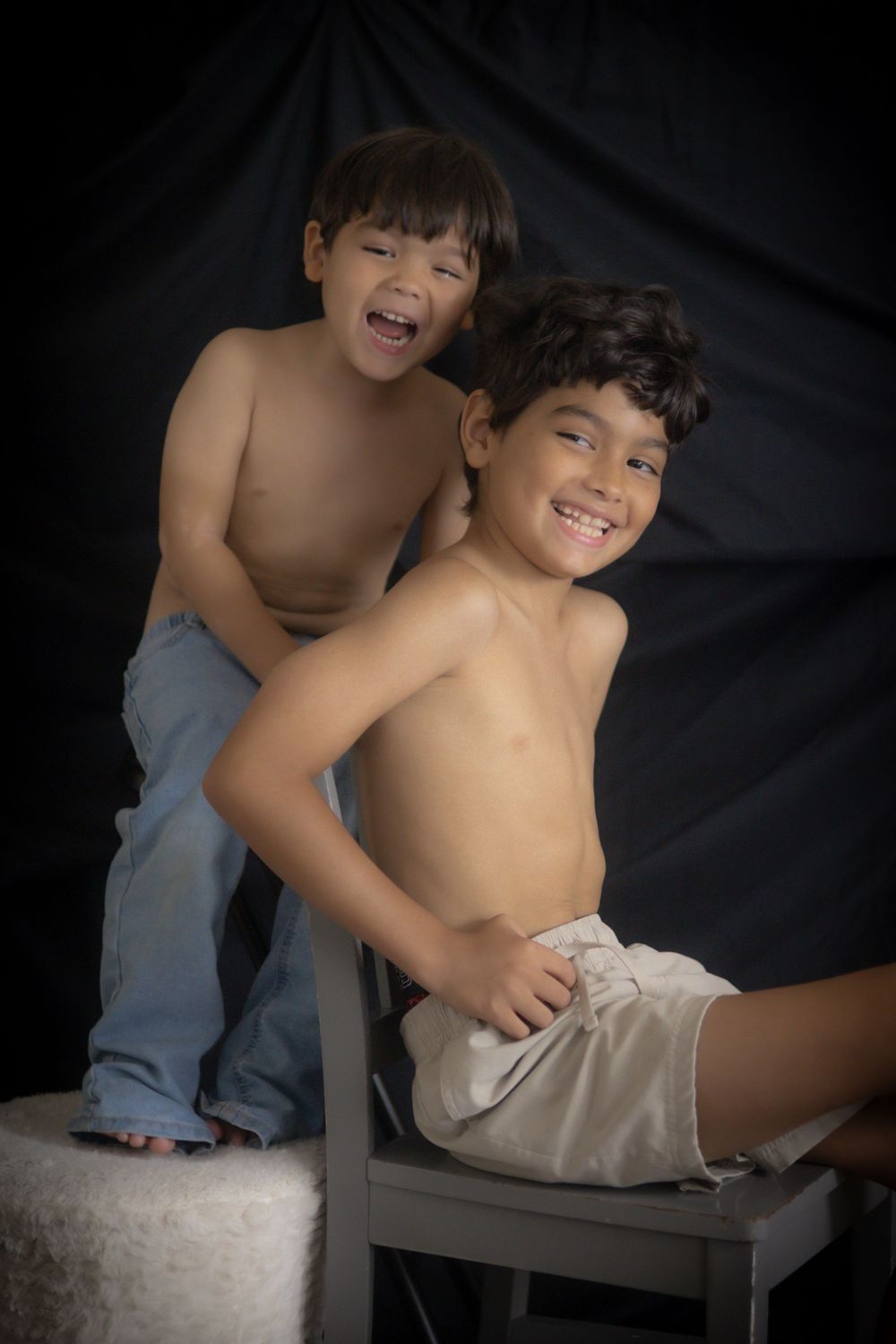 Two shirtless boys laughing and posing together against a dark backdrop in a studio portrait.