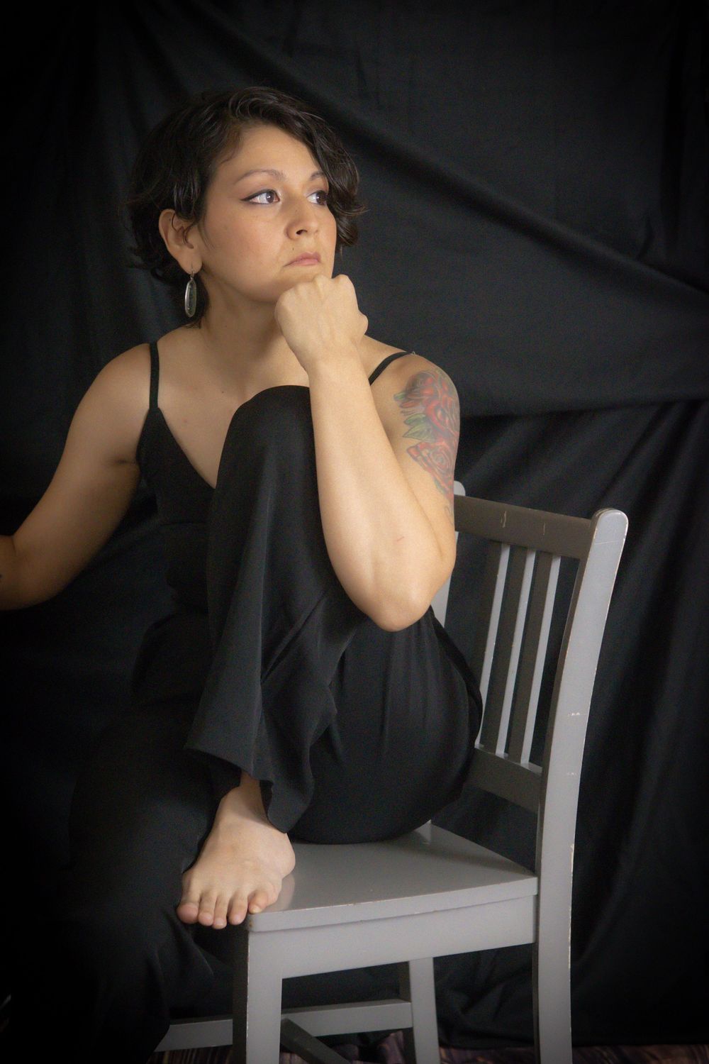 Woman in black jumpsuit with tattoo sits on white chair, posing thoughtfully against dark backdrop.