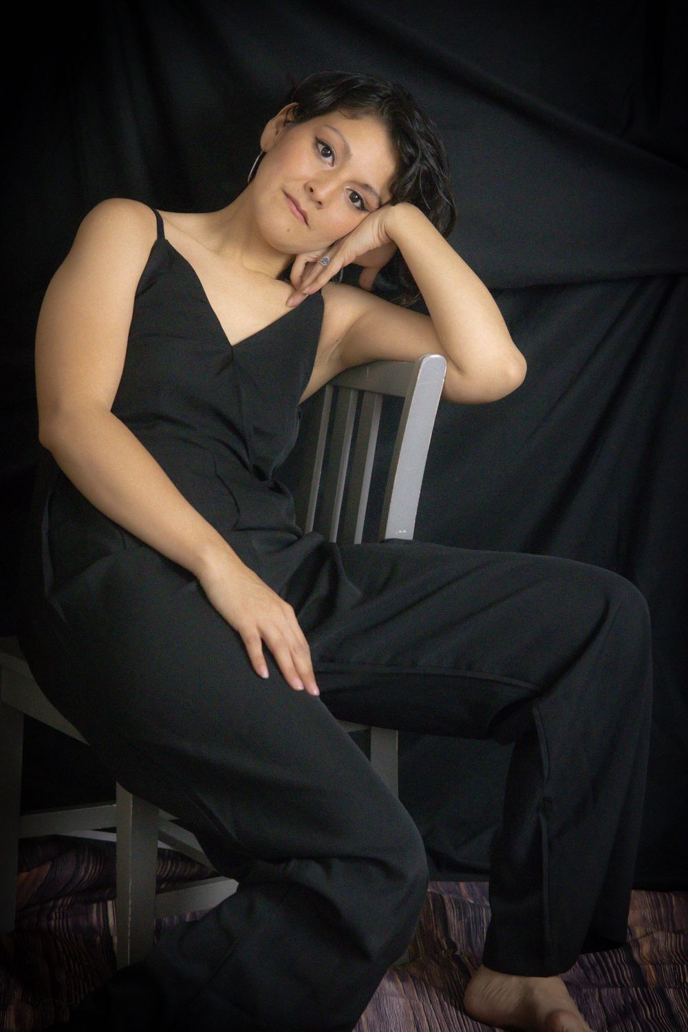 Young woman in black jumpsuit sitting relaxed on a chair against dark backdrop in a studio portrait.