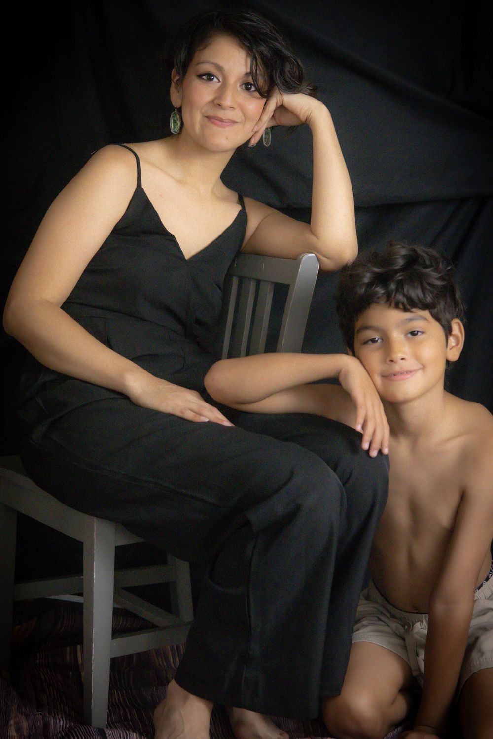 A woman in black jumpsuit sits on a chair beside a smiling shirtless boy against a dark background.