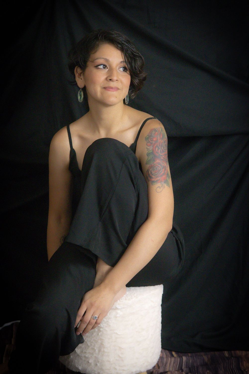 Young woman in black jumpsuit with rose tattoo on arm sits gracefully against dark fabric backdrop.