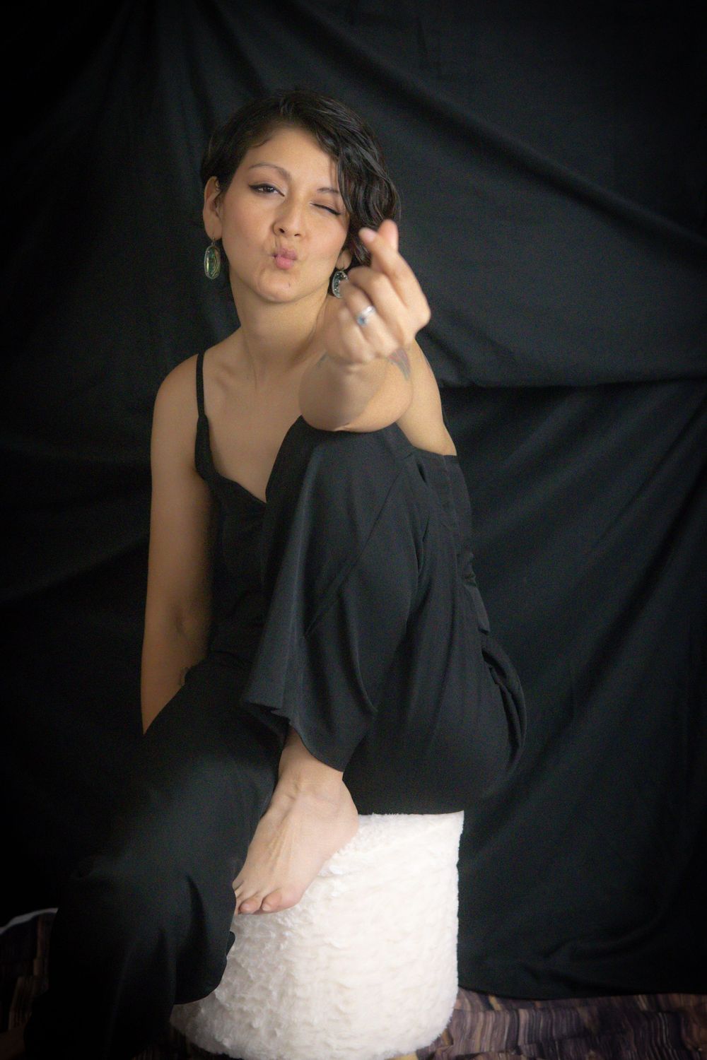 Woman in black jumpsuit sitting on white stool, blowing a kiss toward camera against dark backdrop.