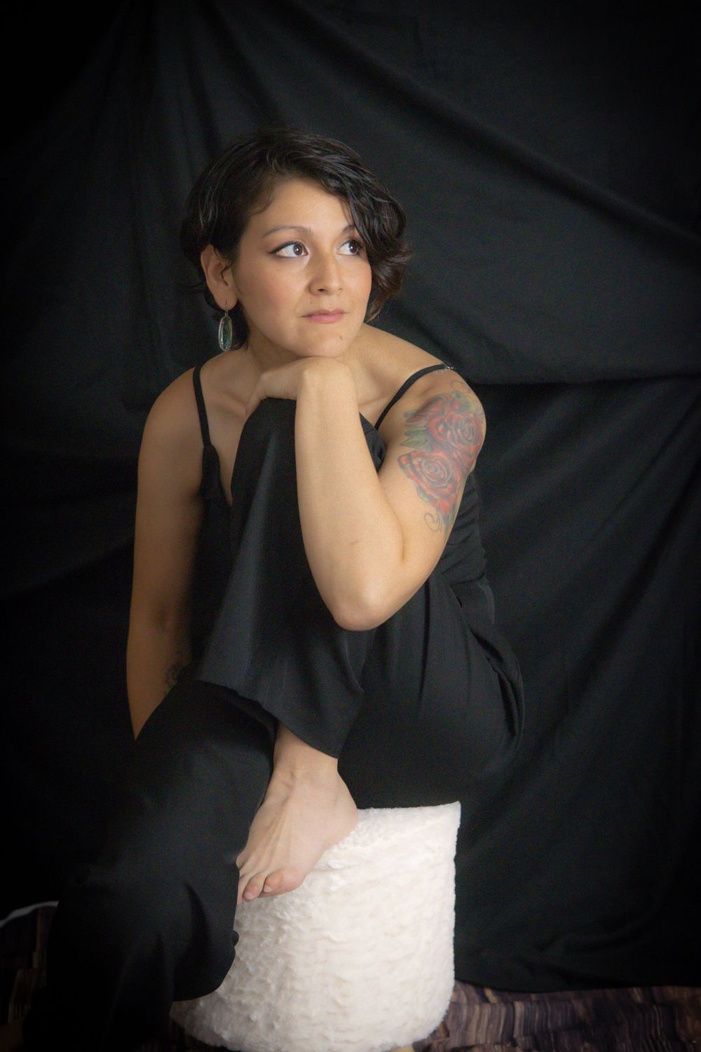 Woman in black jumpsuit with arm tattoo sits on white stool against dark backdrop, gazing thoughtfully to the side.