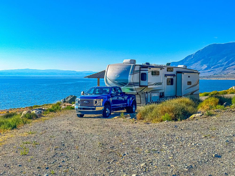 A truck and fifth-wheel RV are parked on a gravel shore beside a vivid blue mountain lake.
