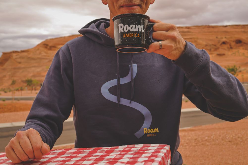 Person sipping from Roam America mug wearing navy hoodie at a red checkered picnic table.