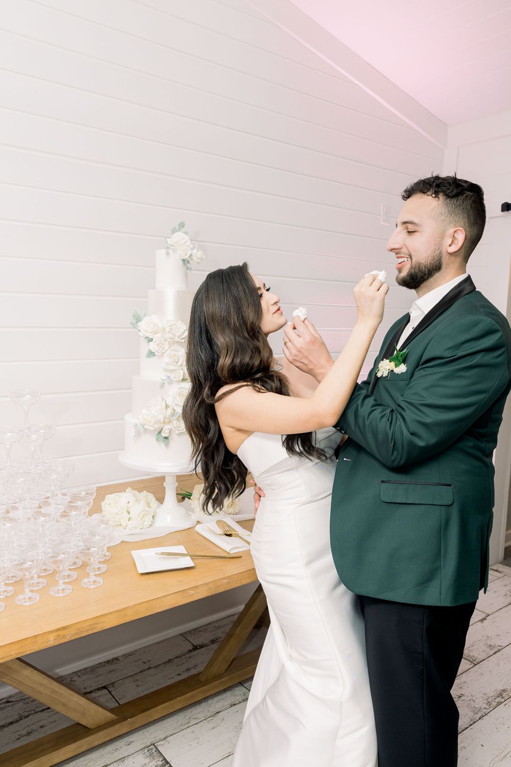 Playful moment of feeding wedding cake between the bride in white and groom in green.