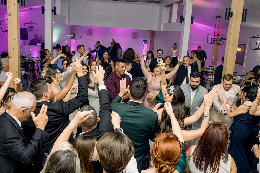 Energetic wedding reception dance floor packed with guests under purple and pink lighting.