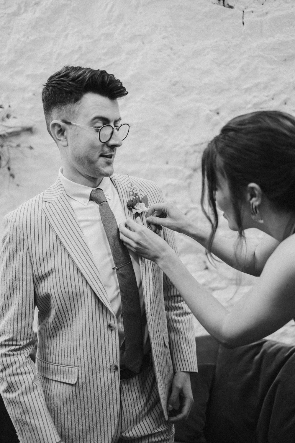 A person in a pinstripe suit and glasses has a boutonniere pinned to their lapel.