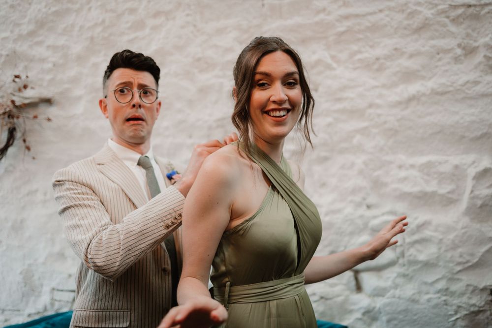A couple in formal attire poses playfully against a white wall.