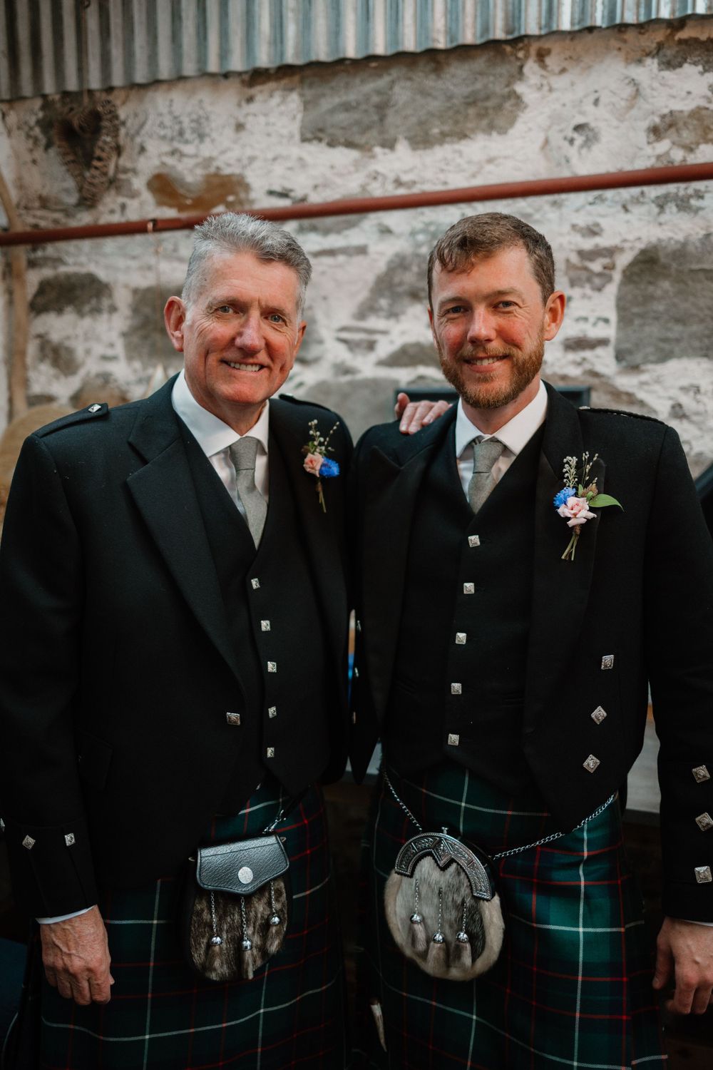 Two men in traditional Scottish kilts and formal jackets pose together against a stone wall.
