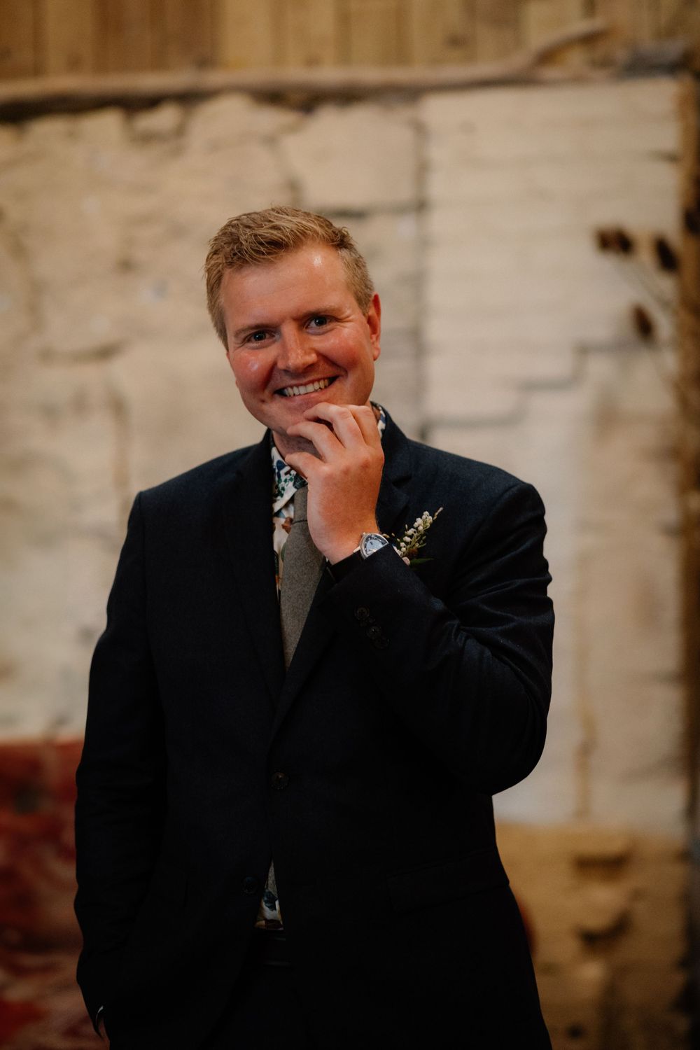 Wedding portrait of a person in a dark suit with a floral boutonniere smiling against a rustic wall.