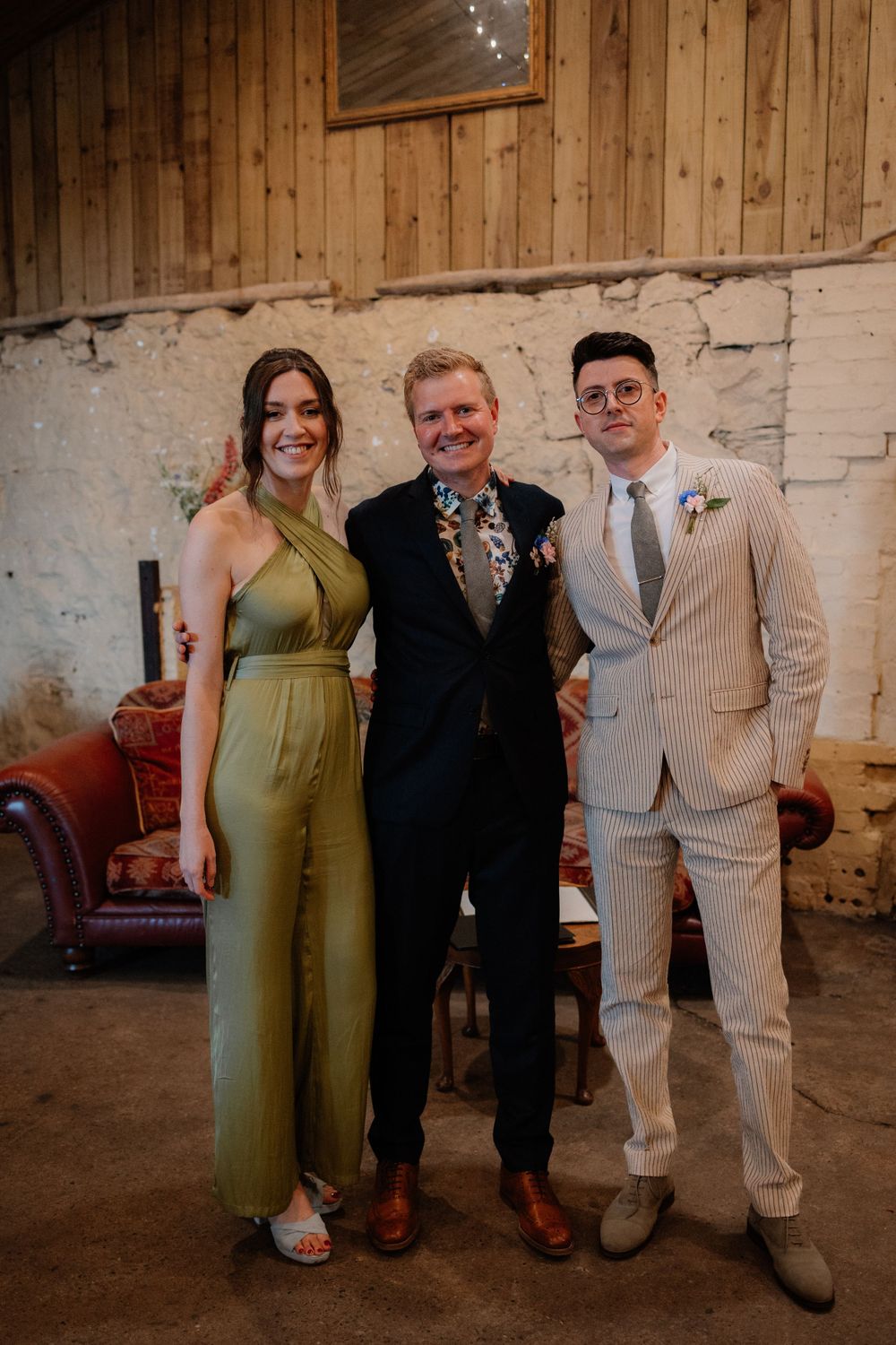 Three people pose together at a wedding wearing stylish outfits including a jumpsuit and pinstripe suit.