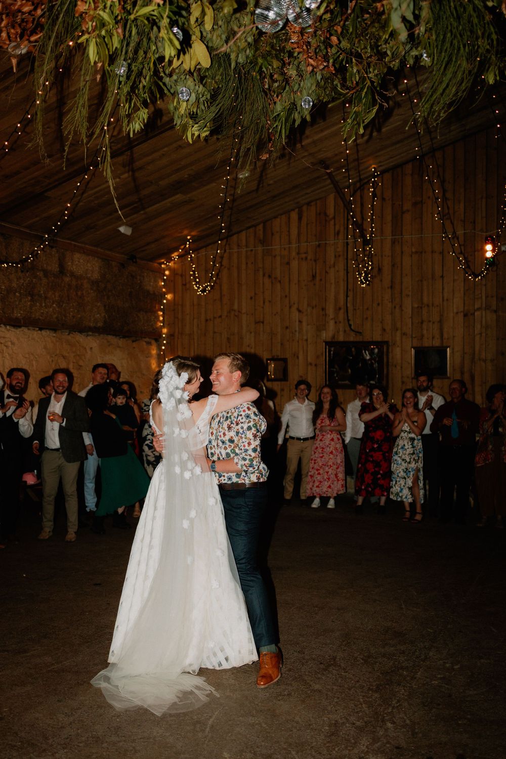 Romantic dance moment in rustic barn venue decorated with greenery and twinkling lights.