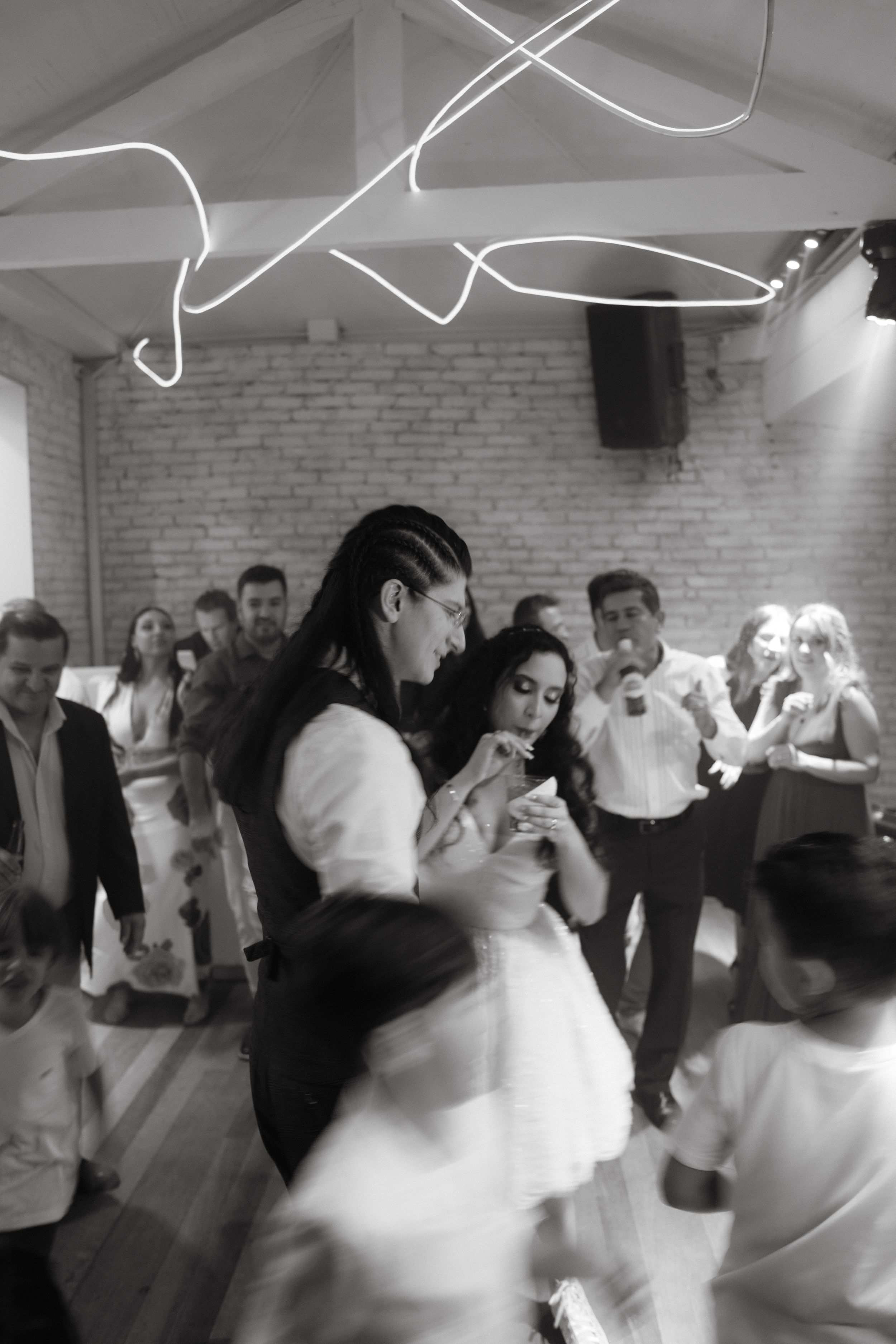 Wedding reception guests dancing energetically on a crowded dance floor with white brick walls and creative lighting.