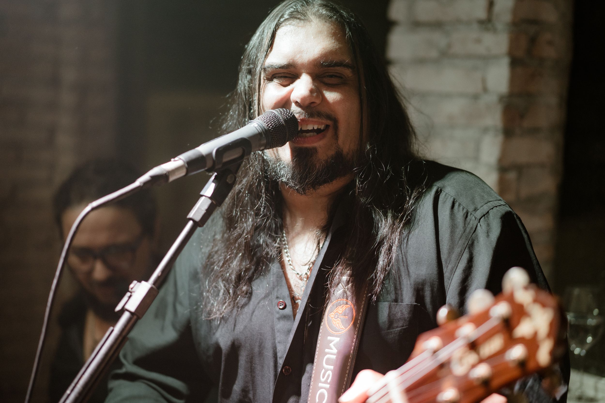 Musician performing with long dark hair singing into microphone while playing acoustic guitar on stage.