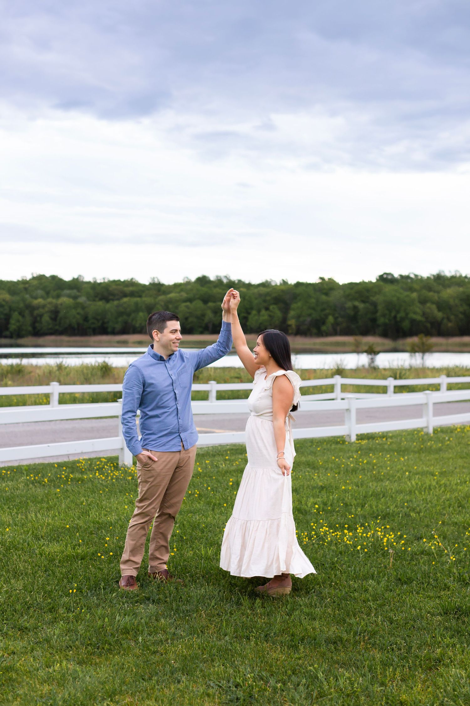 A couple holding hands and dancing in a grassy field with a white fence and lake behind them.