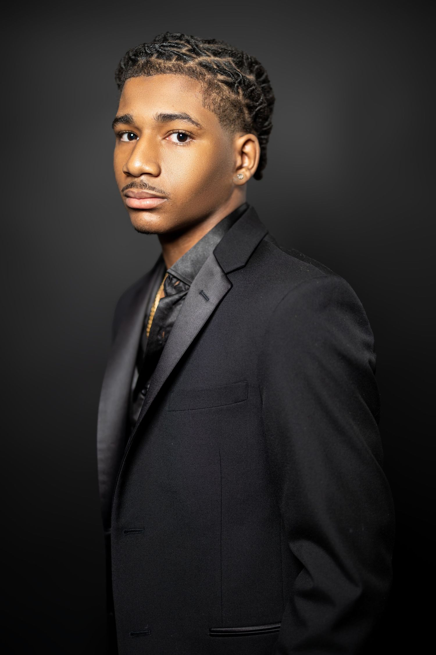 Studio portrait of a young man wearing a black suit and braided hairstyle against dark background.