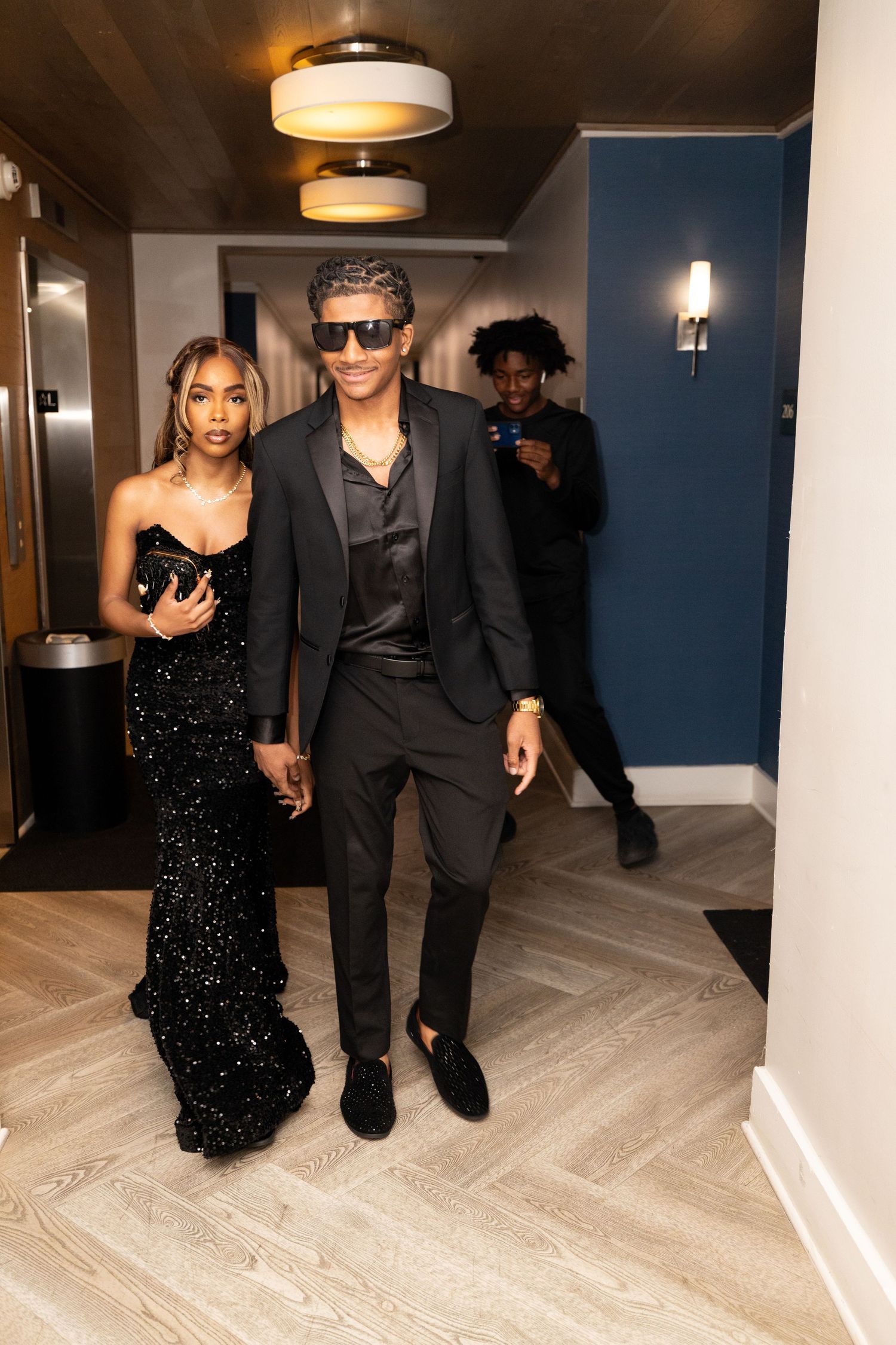 Stylish prom couple in hallway wearing black sequin gown and suit with sunglasses.