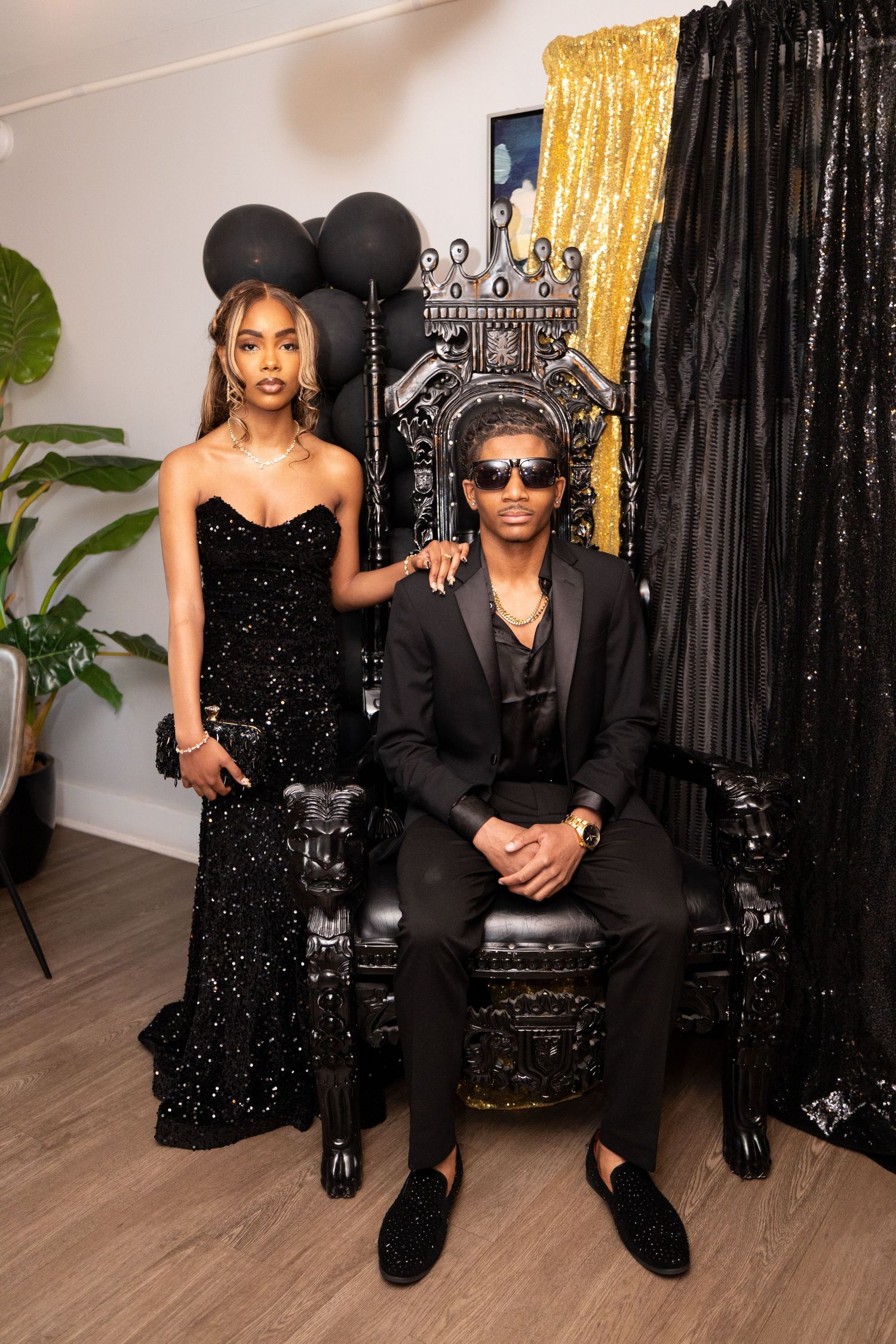 Formal couple posing by decorative throne chair with gold and black backdrop.