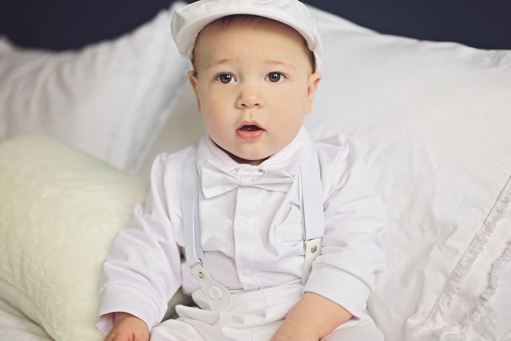 Adorable baby boy in white christening outfit with bow tie and cap sitting on white bedding.