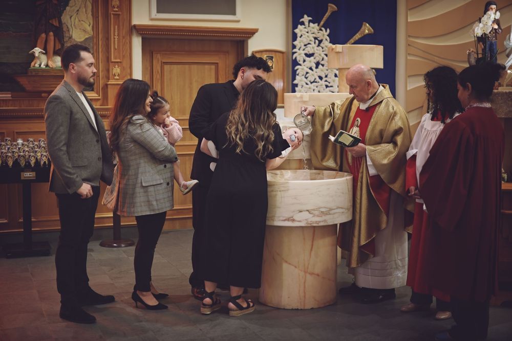 Catholic baptism ceremony at church font with priest in gold vestments and family gathered around.