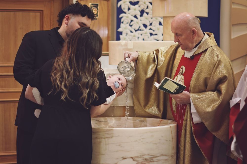 Priest performing a baptism ceremony, pouring holy water while couple watches at a church font.