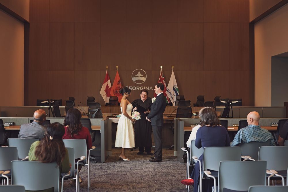 Couple exchanges wedding vows at Toronto City Hall ceremony with guests seated in council chamber.