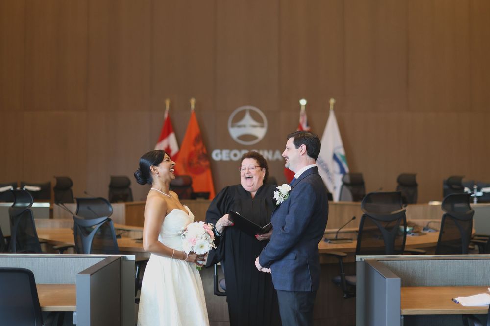 Couple exchanges wedding vows with officiant in a formal council chamber with flags in the background.