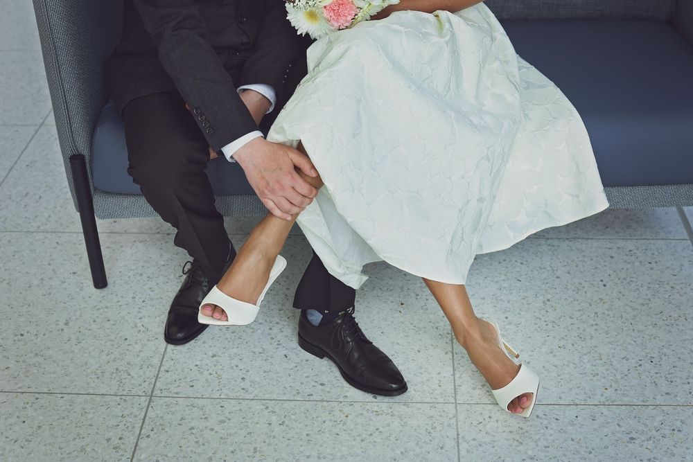 Bride and groom sitting together, showing off wedding shoes, white heels and black dress shoes, with bouquet.