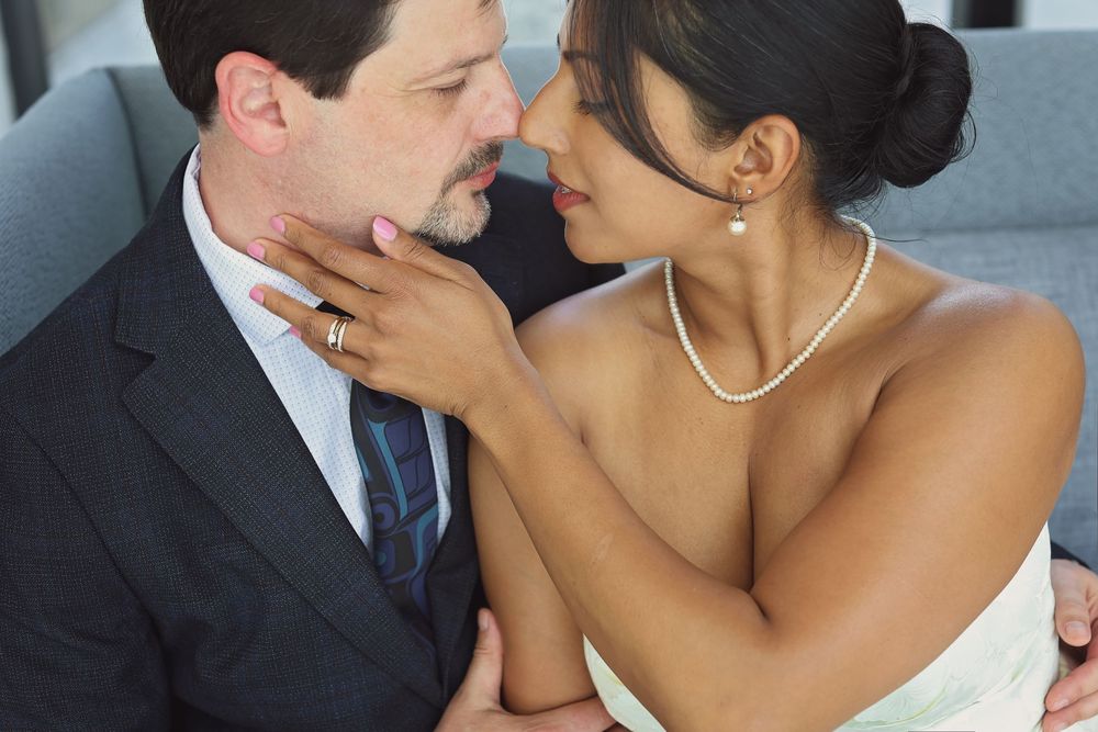 Bride and groom share an intimate moment, with bride in strapless gown and pearl necklace touching grooms face tenderly.