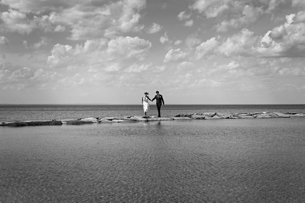Black and white photo of a couple holding hands on a sandy beach path with calm ocean and cloudy sky.