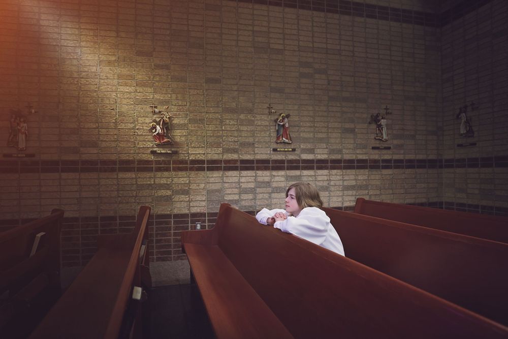 Person sitting alone in church pew, praying beneath religious statues on brick wall.