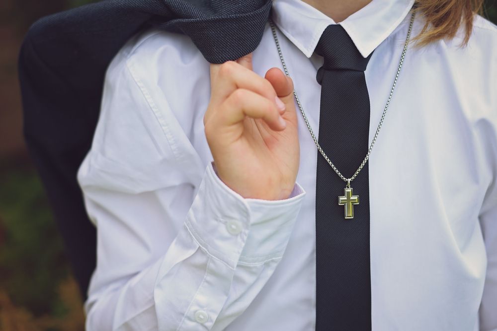 Person in white shirt and black tie wearing a silver cross necklace, holding a dark blazer over their shoulder.