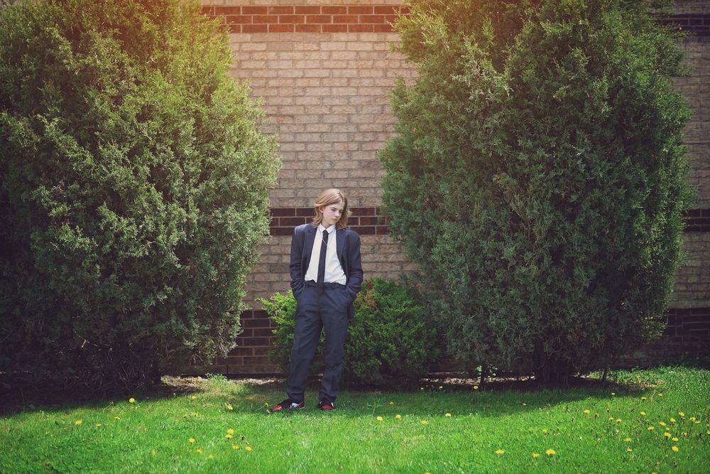 Man in suit standing between tall green shrubs against a brick wall on a green lawn.