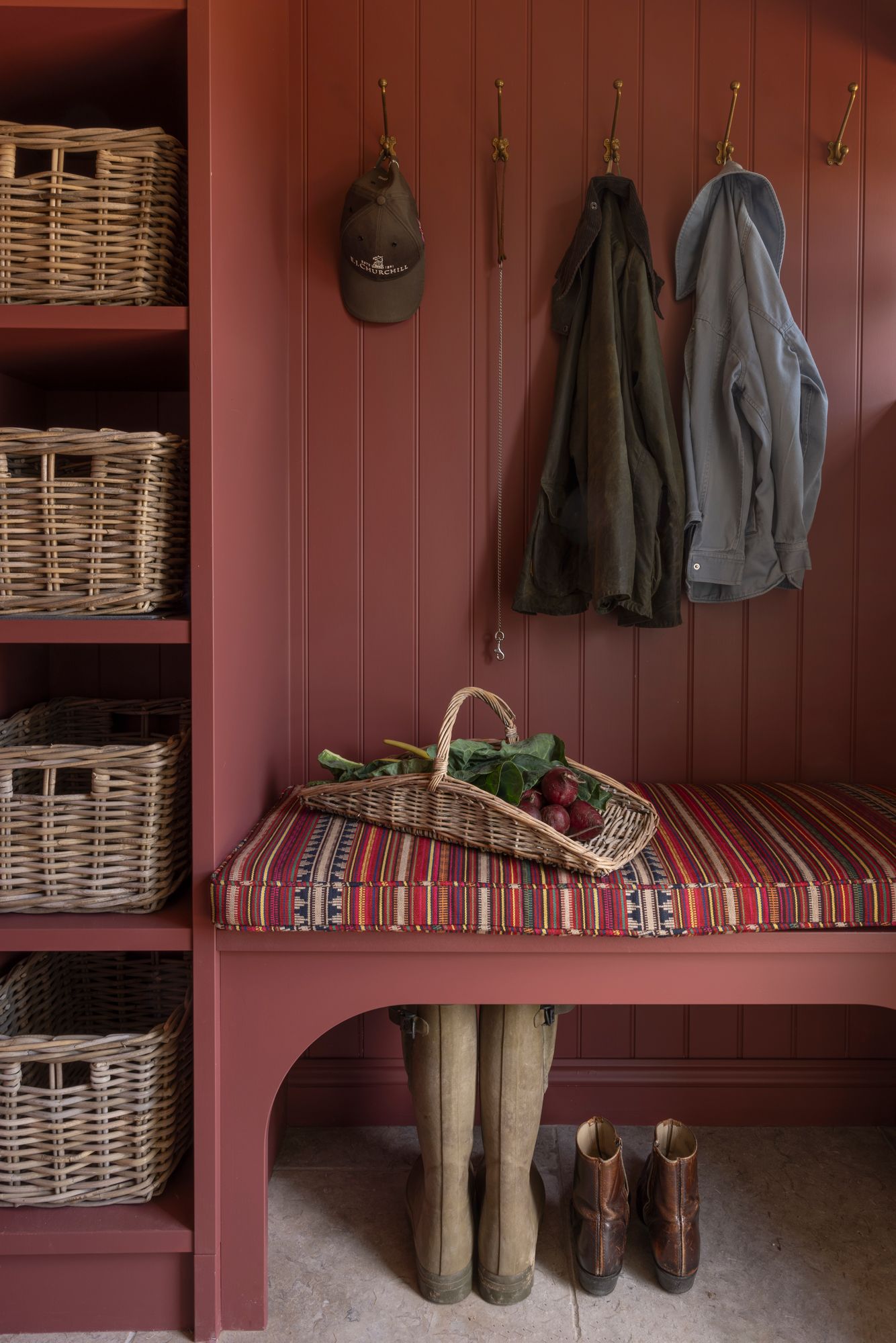 Rustic mudroom with pink walls features white wicker storage baskets, hanging coats, and rubber boots lined up below.