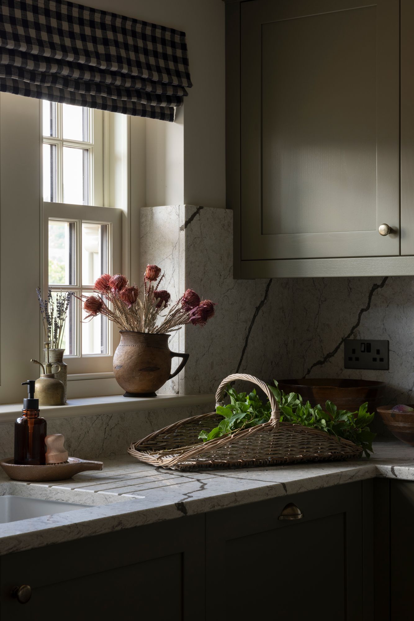 Dark kitchen interior with checkered window valance, marble countertop, and moody natural lighting through window.